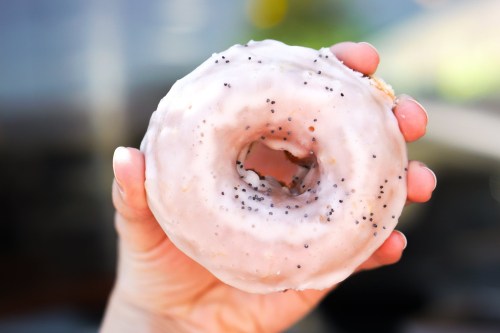 A hand holding a glazed donut topped with poppy seeds against a blurred background.