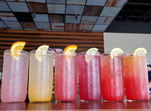 A row of six colorful drinks in mason jars, each garnished with citrus slices, on a wooden table.