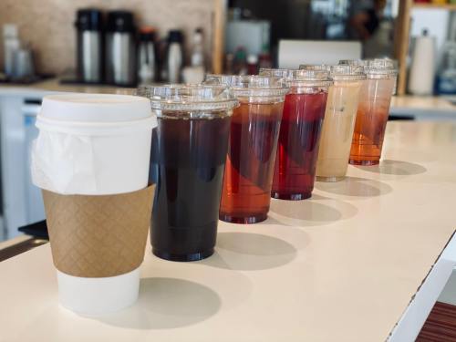 A row of drinks on a counter, including a white coffee cup and several clear cups with various iced beverages.