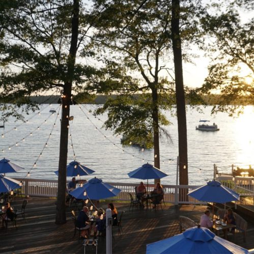 A lakeside restaurant with blue umbrellas, diners enjoying the view, and a boat on the water at sunset.