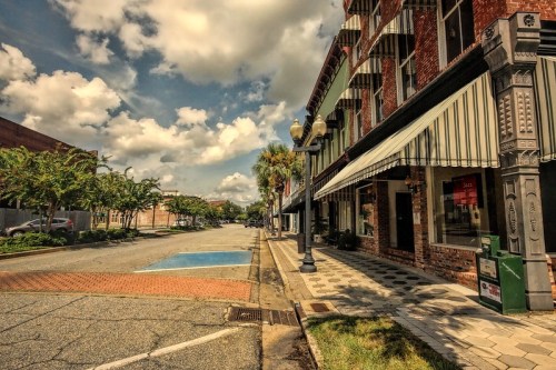 A quiet street scene featuring brick buildings, palm trees, and a partly cloudy sky.
