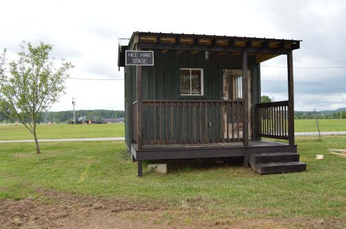 A small green wooden building with a porch, labeled "Hee Haw Stage," set in a grassy area with a cloudy sky.