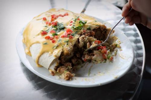 A large burrito topped with cheese, diced tomatoes, and cilantro, with a fork digging into it on a silver plate.