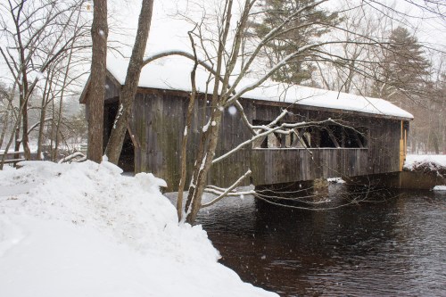 A snow-covered wooden covered bridge spans a river, surrounded by trees in a winter landscape. Snowflakes are falling.
