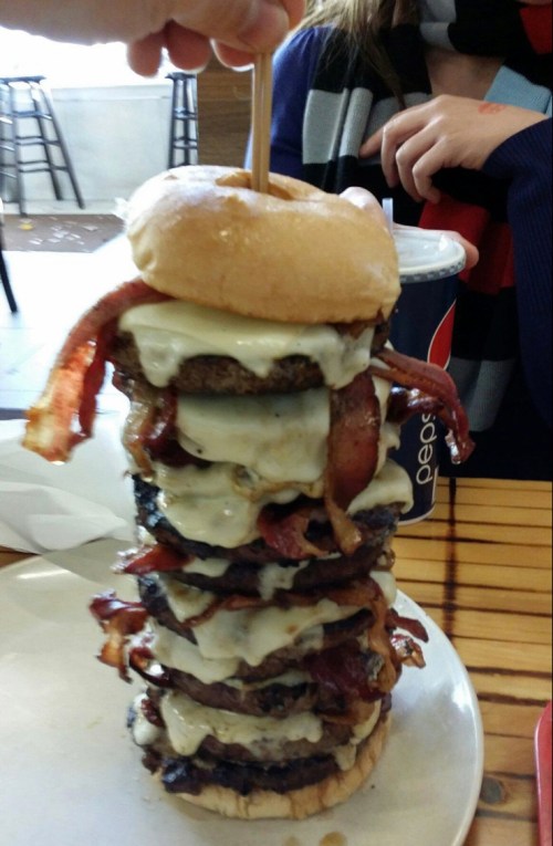 A towering burger stacked with multiple patties, cheese, and bacon, topped with a skewer, alongside a soda.