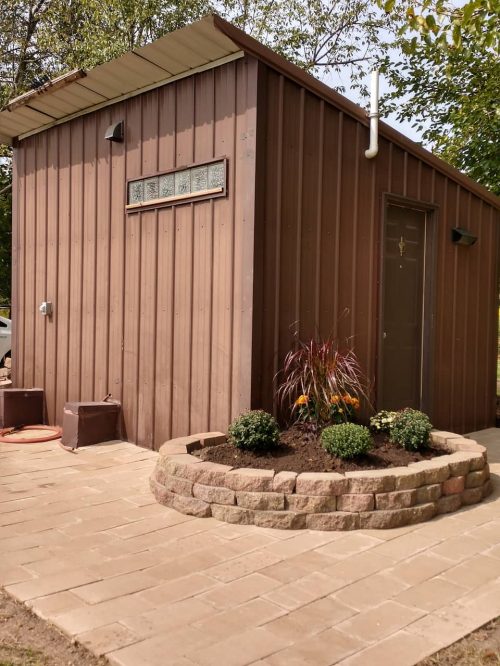 A small brown metal shed with a landscaped flower bed and stone border, surrounded by greenery and a paved pathway.