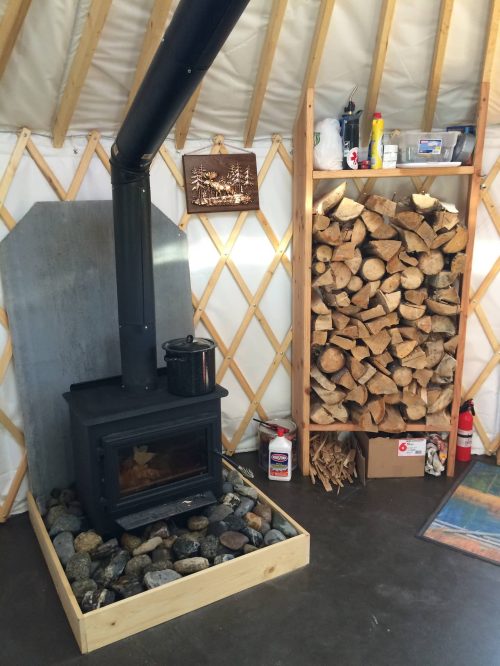Cozy interior of a yurt featuring a wood stove, stacked firewood, and a stone base.