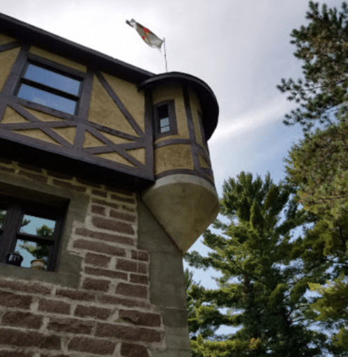 A unique building with a round turret and a flag on top, surrounded by tall trees and a cloudy sky.