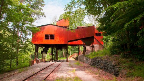 A red industrial building on stilts surrounded by trees and a gravel path, with old railway tracks nearby.
