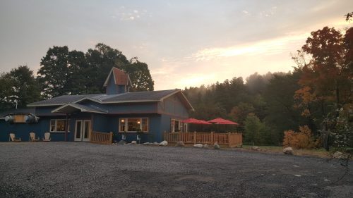 A blue house with a sloped roof, surrounded by trees, features red umbrellas on a patio at sunrise.