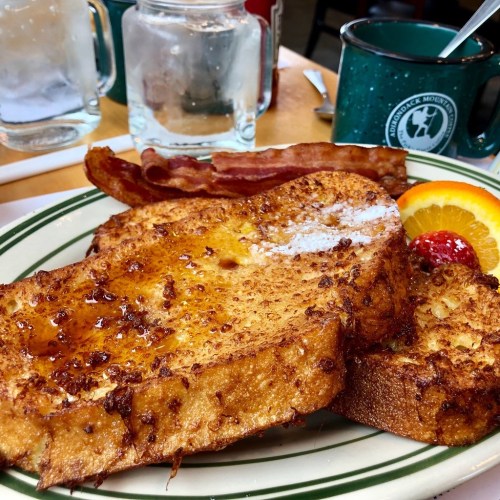 A plate of French toast topped with syrup, served with bacon and a slice of orange and cherry garnish.