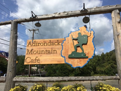 Sign for Adirondack Mountain Café featuring a hiker silhouette and a map outline, surrounded by flowers and blue sky.