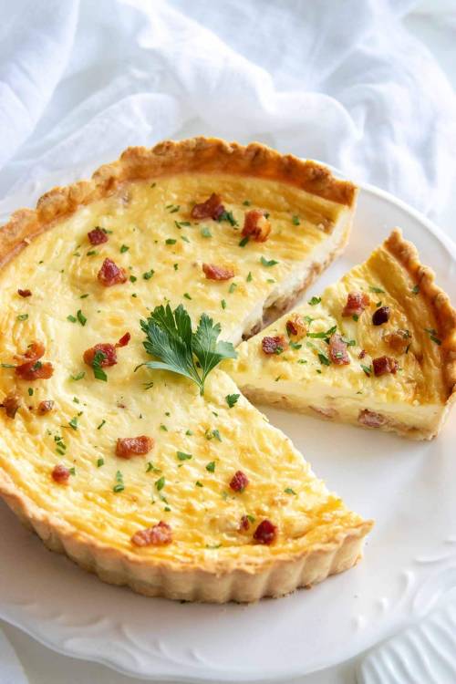 A savory quiche with a golden crust, filled with cheese and bacon, garnished with parsley, with a slice removed.