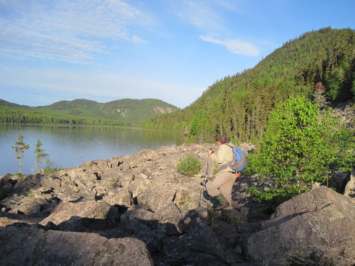 A hiker navigates rocky terrain by a serene lake, surrounded by lush green forests and mountains under a clear blue sky.