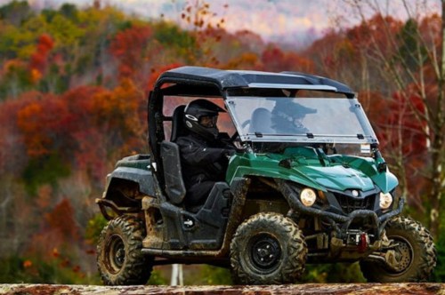 A green all-terrain vehicle drives over a log, surrounded by vibrant autumn foliage.
