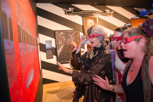 Three women wearing 3D glasses admire a vibrant artwork in a gallery, expressing excitement and engagement.