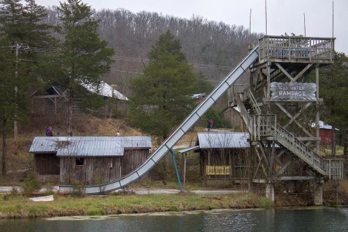 A tall water slide next to an old wooden building, surrounded by trees and a calm body of water.