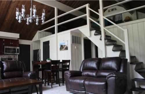 Cozy interior of a cabin featuring leather sofas, a dining area, and a staircase under a wooden ceiling.