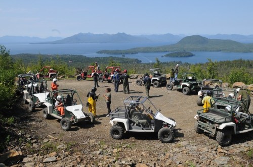 A group of people gathered around off-road vehicles on a scenic overlook with a lake and mountains in the background.