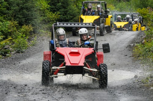 Two people drive a red off-road vehicle through a muddy trail, with yellow buggies following behind in a lush green setting.