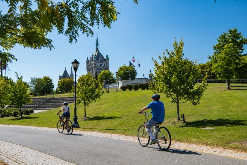 Two cyclists ride along a path in a park, with a historic building and trees in the background under a clear blue sky.