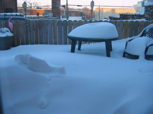 A snowy backyard scene with a covered table, a chair, and a pink flamingo partially buried in snow.
