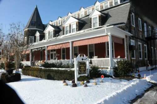A charming inn with a peaked roof, surrounded by snow and greenery, featuring a welcoming porch and sign.