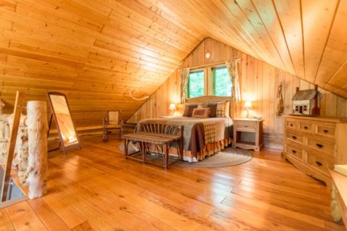 Cozy attic bedroom with wooden walls, a bed, seating area, and warm lighting, surrounded by nature.