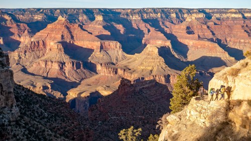 Hikers traverse a rocky path along the Grand Canyon, showcasing its layered red and brown cliffs in the background.