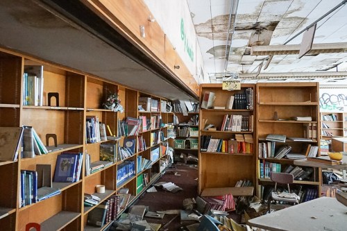 Abandoned library with disorganized bookshelves, scattered books, and signs of decay on the ceiling.