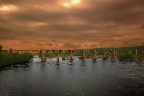 A stone bridge with multiple arches spans a river under a dramatic, cloudy sunset sky.