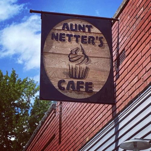 Sign for Aunt Netter's Cafe featuring a cupcake design, hanging from a brick building under a blue sky.