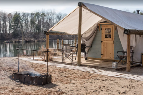 A cozy glamping tent by a river, with a fire pit and chairs on a sandy area, surrounded by trees.