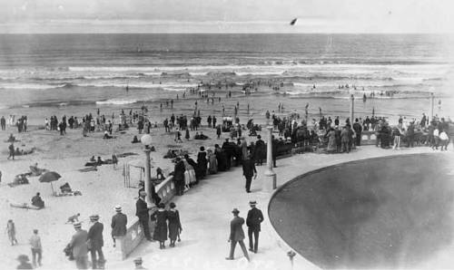 A historic beach scene with crowds enjoying the shore, waves, and promenade on a sunny day.