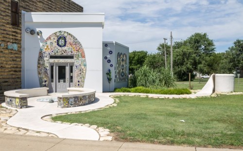 A colorful mosaic-covered building with a curved pathway and green grass, set against a partly cloudy sky.