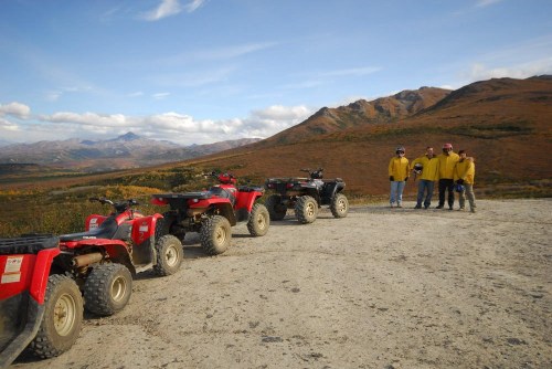 Four people in yellow jackets stand beside parked ATVs on a dirt path with mountains and autumn foliage in the background.