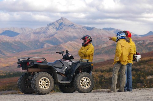 Three people in yellow jackets and helmets stand near an ATV, pointing towards a mountain landscape in the background.