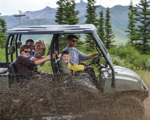 A group of four people in an off-road vehicle splashes through mud, surrounded by trees and mountains.