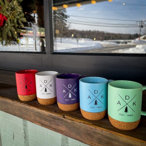 Five colorful mugs with cork bases, featuring a logo, lined up on a wooden ledge against a snowy backdrop.