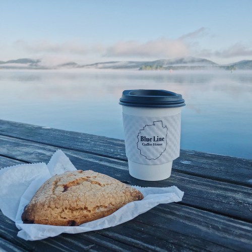 A coffee cup and a scone sit on a wooden table by a calm lake, with misty mountains in the background.