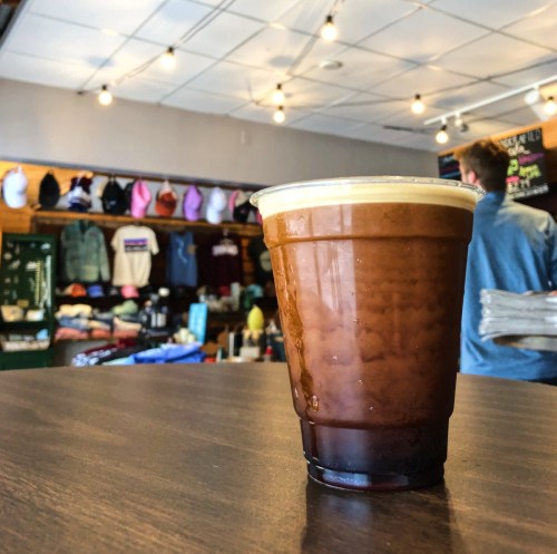 A cold coffee drink sits on a table in a cozy shop with clothing and hats displayed in the background.