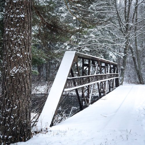 A snow-covered bridge spans a quiet river, surrounded by trees in a serene winter landscape.