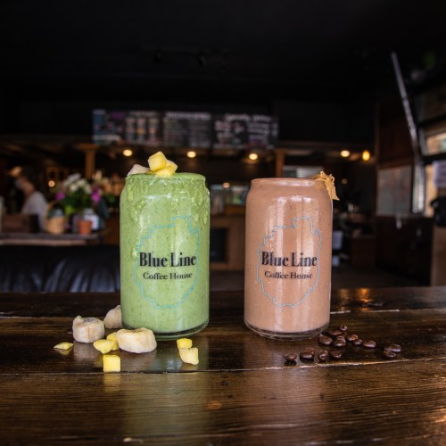 Two smoothie jars from Blue Line Coffee House: one green with fruit, the other brown with a nutty topping, on a wooden table.