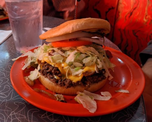 A close-up of a cheeseburger with lettuce, tomato, and onion on a red plate, with a glass of water in the background.