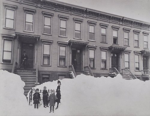 Black and white photo of children playing in deep snow in front of a row of brownstone houses.