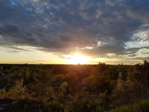 A sunset over a lush landscape, with clouds scattered across the sky and a warm glow illuminating the horizon.