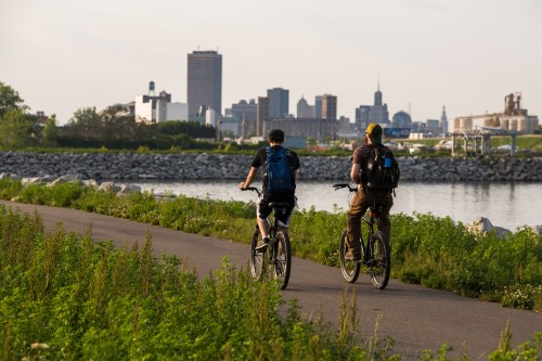 Two cyclists ride along a path by the water, with a city skyline in the background on a sunny day.