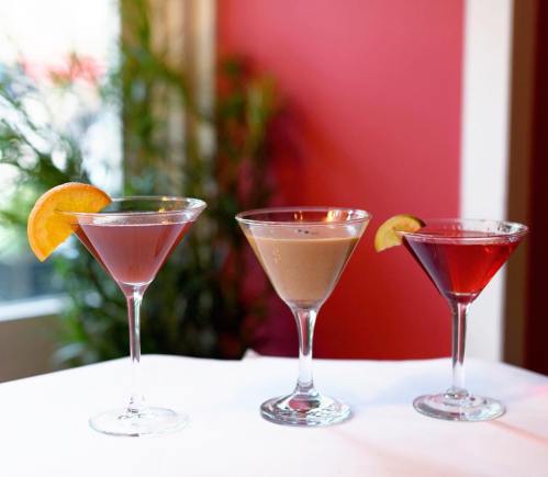 Three colorful cocktails in elegant glasses, garnished with orange and lime slices, set against a vibrant red background.