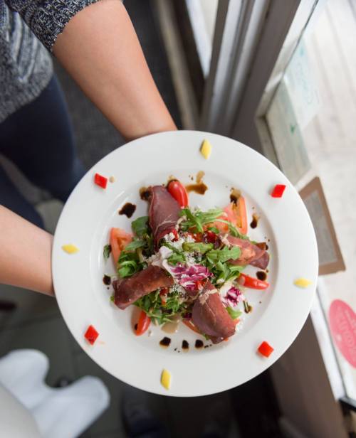 A person holds a plate of colorful salad with greens, tomatoes, and drizzled balsamic glaze, viewed from above.