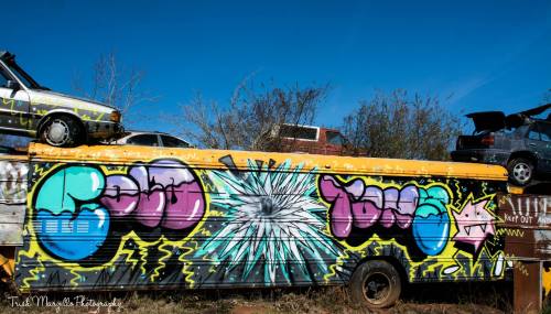 Colorful graffiti on a yellow trailer, featuring bold text and a starburst design, with junked cars in the background.